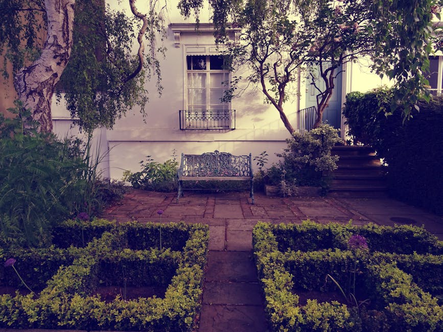 A backyard garden scene featuring neatly trimmed lawn areas bordered by rectangular garden beds filled with a variety of plants and shrubs. In the foreground, wet soil and mulch surround small floweri