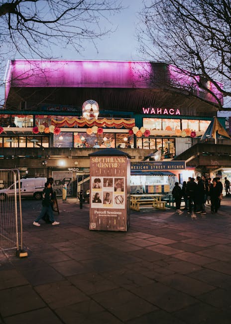 An illuminated urban scene capturing a lively marketplace or outdoor dining area at dusk, with a large, modern building in the background featuring a vibrant pink and purple LED-lit roof. The foregrou
